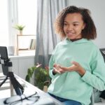Pretty teenage girl in casualwear sitting by table in bedroom and recording video report for seminar while looking in smarthone camera